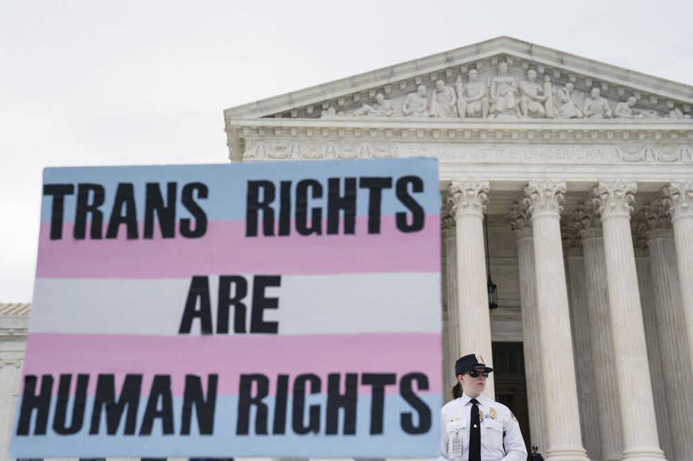 Picture of a Washington, DC building with a security guard standing in front. In front of the security guard is a sign with the trans flag that says "Trans Rights are Human Rights."