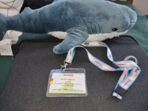 Picture of Noah Adams' EPATH name badge on a lanyard sitting on a grey background. There is a stuff shark (Blahaj from Ikea) above.